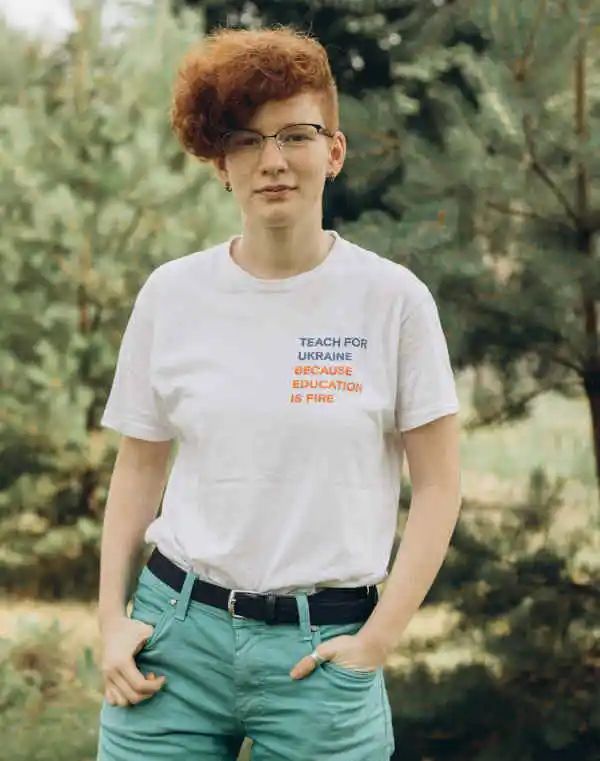 A photo of Yulia. She has red curly hair and glasses and she is wearing a tshirt that says “Teach for Ukraine.”