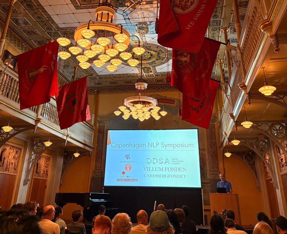 Desmond at the podium in front of a slide presentation with the name of the event. The room is formal and has big chandeliers and flags.