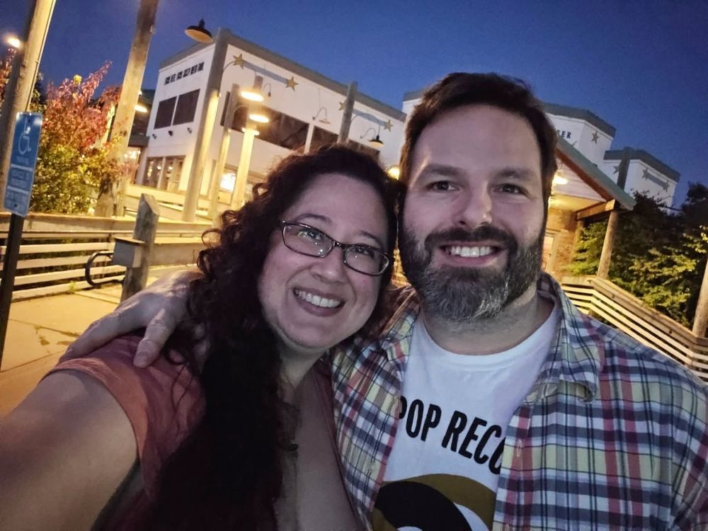Smiling couple stand in front of a building in the evening. Both have dark brown hair. She wears glasses and he has on a Sub Pop shirt under a plaid shirt. 