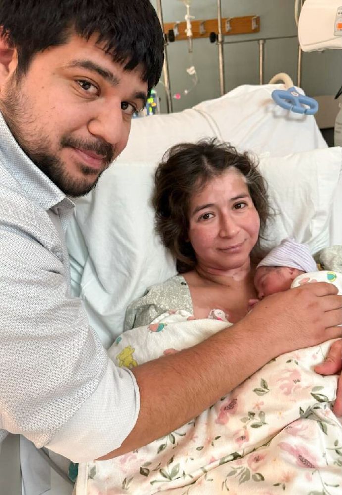 Leo reaches over Vanessa holding newborn Lyla in the hospital bed.