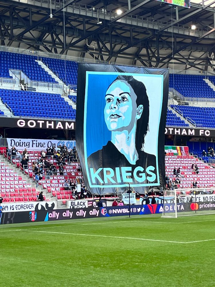 Cartoon portrait of Ali Krieger in varying shades of blue. “Kriegs” in giant capital letters across the bottom.