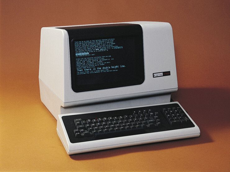 A catalog shot of the VT-100 features a white-on-black monichrome monitor, a black monitor inset in a white case, and a black keyboard frontispiece and keys over a white keyboard case, set in a 70s warm umber backshot. The text, barely visible, features various forms of graphics characters, fonts, double-height lines, and inverse video; the 'digital VT 100' slogan appears case right.