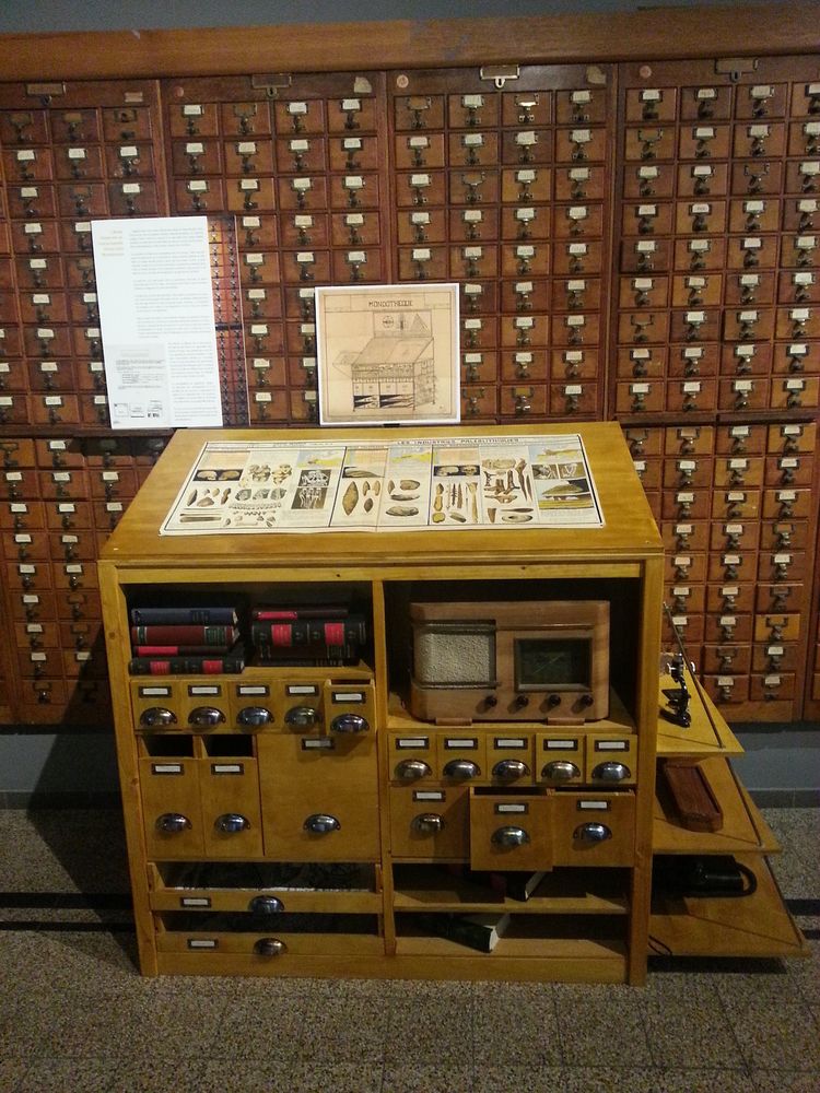 A shelving unit holding a small rough-hewn electric appliance. In comparison to the card catalogues behind it, it's a tiny device, no longer than a desktop radio of the era; documents produced by this 'electric telescope' could be reviewed on the desk itself.