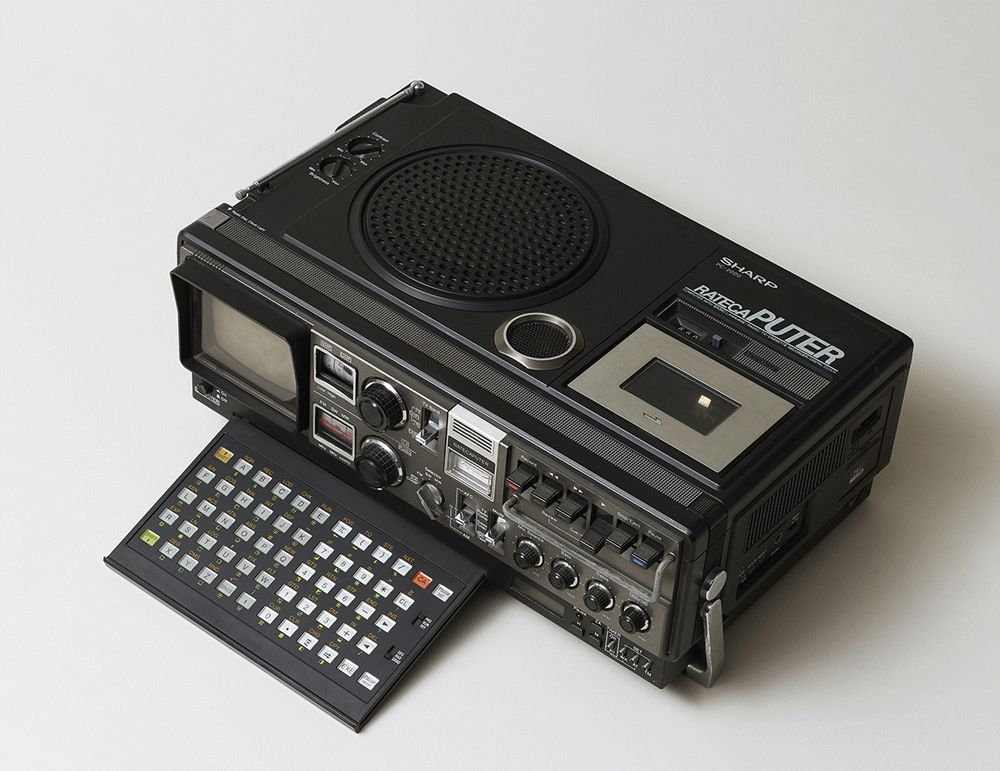 The Sharp Rateca Puter. Up top, left to right: a collapsable TV/radio antenna, brightness and contrast dials, a giant speaker, a smaller speaker, and input for the tapedeck, with the play counters that were more common on the day. Face, left to right: A 4.5 inch CRT (behind a plastic guard), VHF/UHF and radio dials, channel and band selectors (TV/COMP, FM, Shortwave, and Middlewave), the "RATECA PUTER' logo on the battery gauge, AFC switch, mode selector, tape deck controls, audio controls for balance, bass, treble, and volume, and a mollyguard to prevent dials and switches from being mashed (the mollyguard being a signature of Sharp's boomboxes of the time). Face, low: the collapsable carrying handle that doubles as a stand; computer power button; slide-out chiclet keyboard based on Sharp calculators of the day, with number pad; an LED clock, clock controls, alarm controls. Right side: mic jacks, earphone jacks in two different sizes, beat cut switch, fan exhaust, DC and AC power plugs, and a printer port (!). Help I'm trapped in an alt text factory