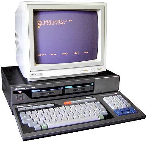 A three-piece computer unit. Up top, the white monitor reads: "MSX BASIC versão 1.1 Br 'G r a d i e n t e' 28014 bytes livres OK" up top, then "color auto goto list run" down below. The computer itself is a grey pizzabox, with black angular highlighting joining the two cartidge slots and the power light. Image bottom, the Expert's keyboard; a full keyboard, it includes multimedia buttons, a blue directional keypad, and the same angular black highlighting as is on the face of the computer proper. A small recess in the computer allows the keyboard to be locked in place, although this seems only to be to hold the keyboard in position, not to store the keyboard.