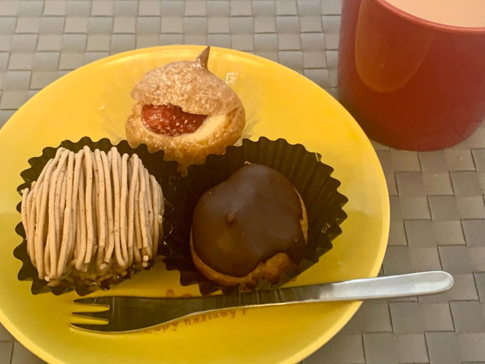 小さいケーキ3種（モンブラン、いちご入りシュークリーム、エクレア）が昔ローソンのおまけでもらったリラックマの直径15cmぐらの黄色い皿に乗っている写真。柳宗里のヒメフォークが添えられ、横に赤いイッタラのマグカップ（大好き）が置いてある。中身はキャラメルミルクティー。下に敷いてるのはグレーのプラスチックっぽい素材のあみあみランチョンマット。確かイケアで買ったやつ。すっきり見えて汚れが落ちやすく安価故に容赦なく使えるので便利。