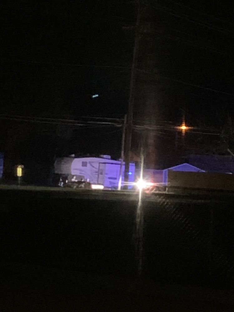 blurry picture of a white rv in the distance with a bright light shining off a cop car while 3-4 police men search a white truck parked near the front of the rv 