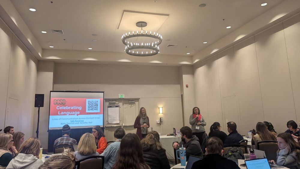 Two presenters at a conference with a "Celebrating Language" slideshow and wipebook displayed. 