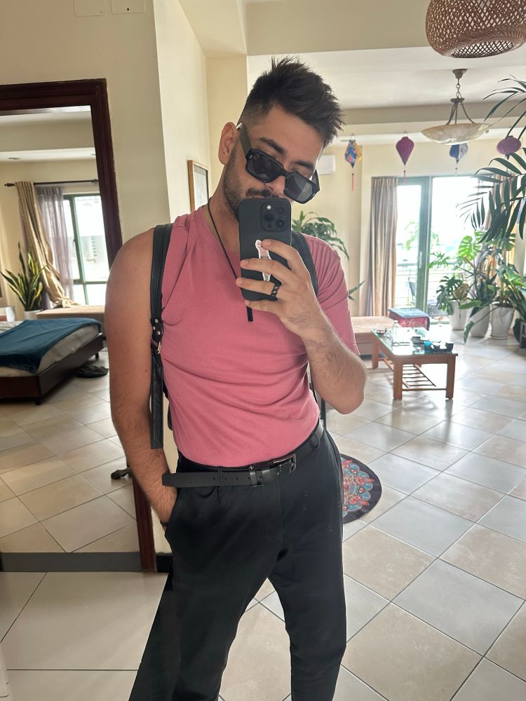 Mirror shot of a confident man in tailored black trousers and a tucked pink tee, radiating quiet confidence and soft-masculine energy.