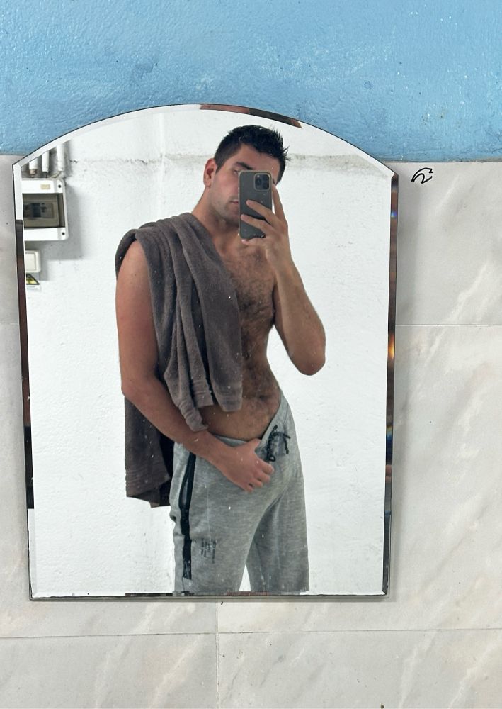 Tall Mediterranean guy half-naked  posing in gray sweatpants posing in front of a mirror with a gray towel covering his left side