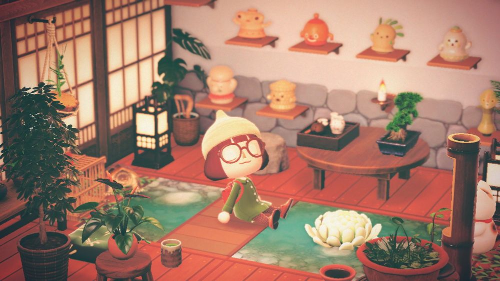 Screenshot of one the rooms in my Animal Crossing: New Horizons house. It’s a very relaxing nature-themed space with warm brown wooden floor and furniture, soft lighting, plants, and a wall full of natural gyroids on shelves. My player character, wearing a cream acorn knit cap and round brown glasses, is sitting on a wooden plank that’s crossing a small indoor garden pond with white lotus flower.