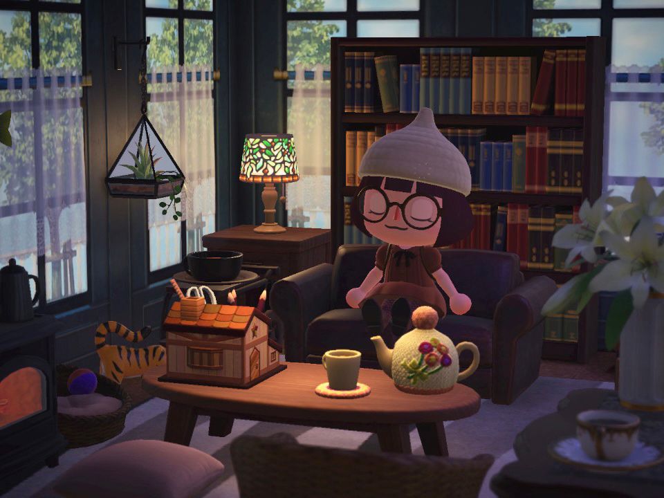 Screenshot of the attic in my Animal Crossing house. It’s a very cozy dimly lit room with antique furniture, a furnace and tall windows. My player character, wearing a cream acorn knit cap, round glasses and cute brown dress, is dozing off on an old leather couch. In front is a low wooden table with a knitted tea cosy and pencil holder shaped like a house.
