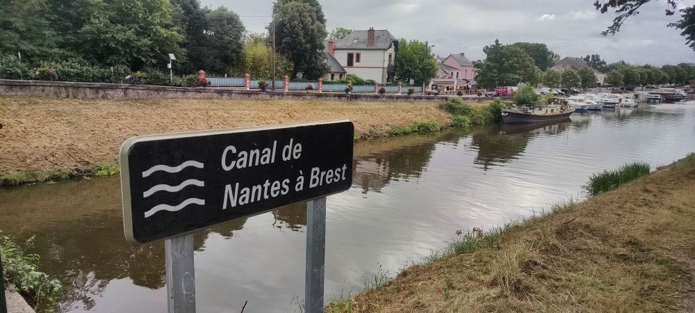 Panneau indiquant "Canal de Nantes à Brest", avec en fond des péniches sur l'eau et quelques maisons