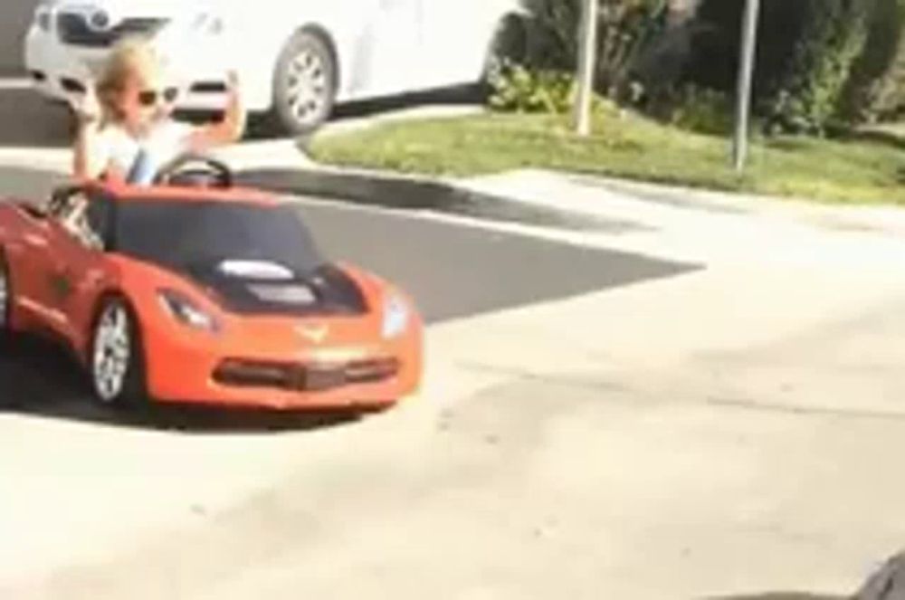 a little girl is driving a red toy car down a street .