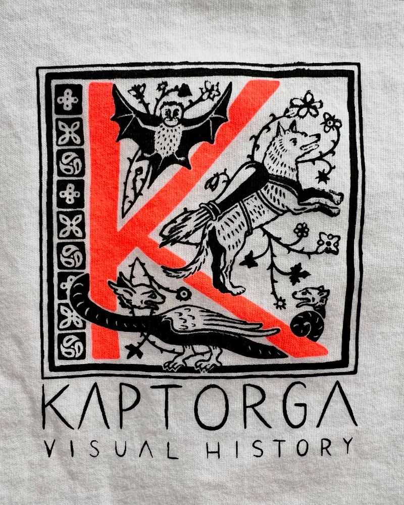 Close-up of a black and neon red print on white jersey fabric, showing a capital K (neon red) with several medieval creatures frolicking around it (black), below it the words „Kaptorga - Visual History“ (black).