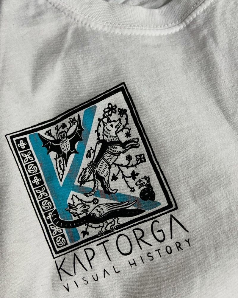 Close-up of a black and swimming pool blue print on white jersey fabric, showing a capital K (blue) with several medieval creatures frolicking around it (black), below it the words „Kaptorga - Visual History“ (black).