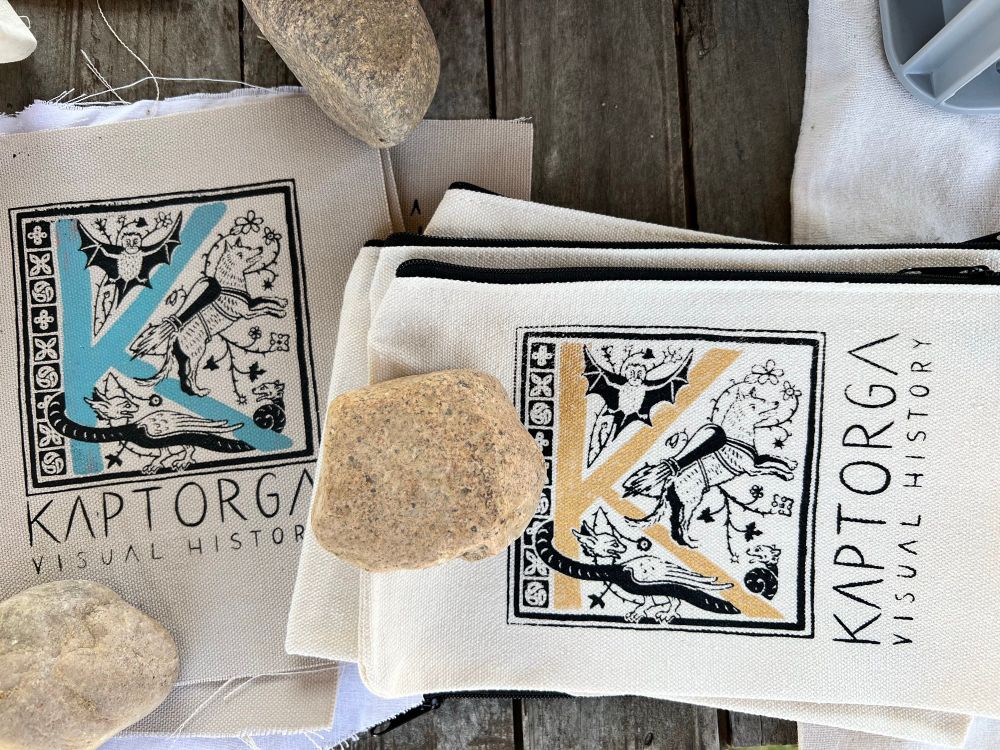 Patches und Reißverschlusstäschchen, die mit einem mittelalterlich anmutenden Initial „K“ nebst dem Schriftzug „Kaptorga - Visual History“ bedruckt sind. Die Artikel liegen auf einem dunklen Holztisch und sind mit Steinen beschwert.