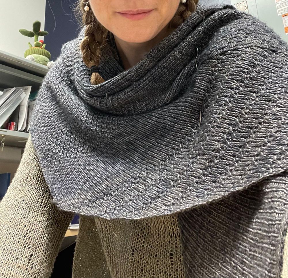 Photo show the torso of a woman wearing a patterned shawl knit from deep blue and grey yarn. Beneath the shawl you can glimpse a grey lacy weight sweater. 