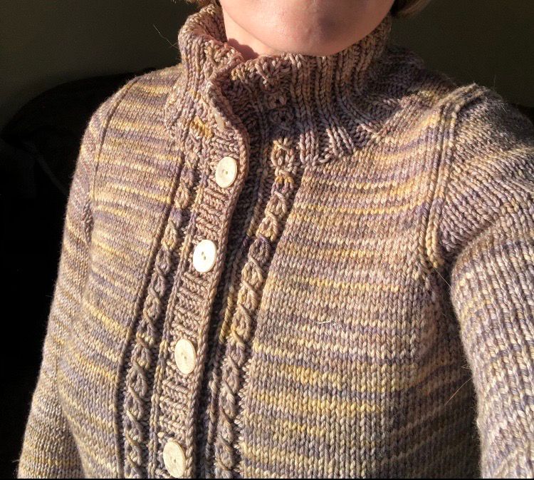 The photo shows the torso of a woman, showcasing a knitted cardigan made from variegated dusty purple to light yellow yarn. The sunlight makes the buttons shine and brings out the mock cable on either side. 
