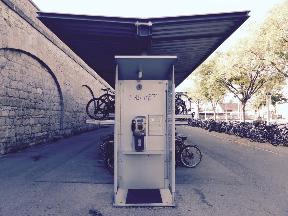 Eine öffentliche Telefonzelle an einem Bahnhof. Links davon ist die Bahnhofsmauer, dahinter und rechts von der Telefonzelle stehen viele Fahrräder. Die Telefonzelle hat keine Tür, und über dem Telefon selbst stehen in schwarzer Handschrift die Worte Call Me.