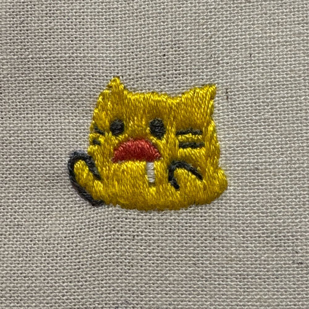 黄色い猫（blobcat）の絵文字の刺繍です。前腕を挙げ、大きく口を開けて涎を垂らしています。かわいい。