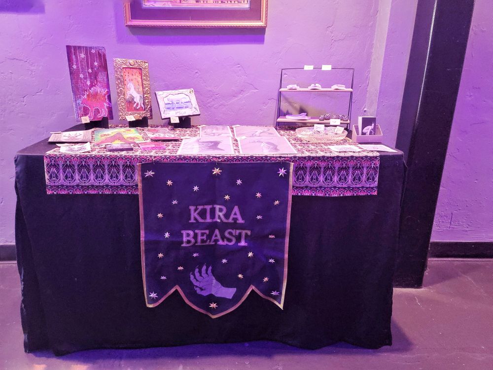 A photo of a table set up to sell art. It has many piece of art sitting in top of it. On the front of the table is a banner hanging that reads:

KIRA
BEAST

The banner is black with a gold border. Below the text, there is an image of a clawed hand. Filling in the negative space on the banner are a variety of silver and gold stars. 
