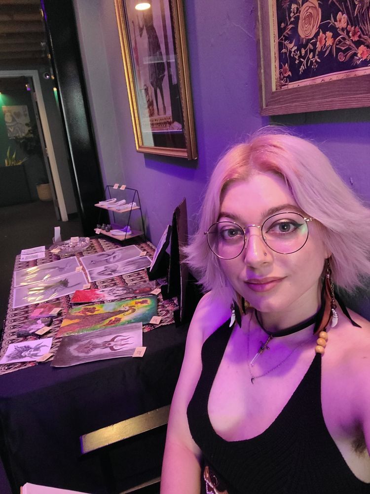 A selfie of a person with pale pink hair sitting to the side of a table set up to sell art. 