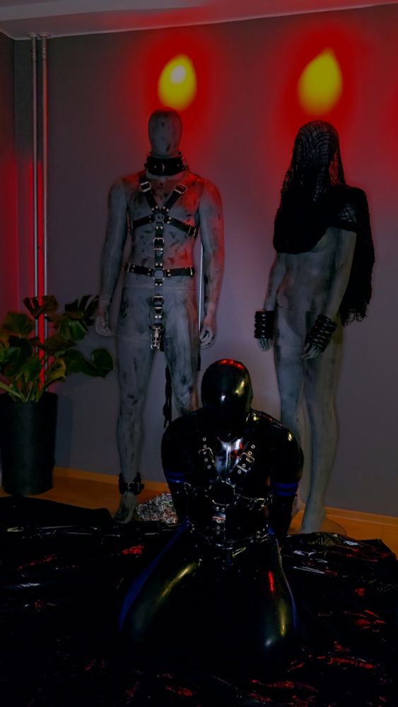 A rubber drone in black latex kneeling in front of two mannequins in BDSM gear.