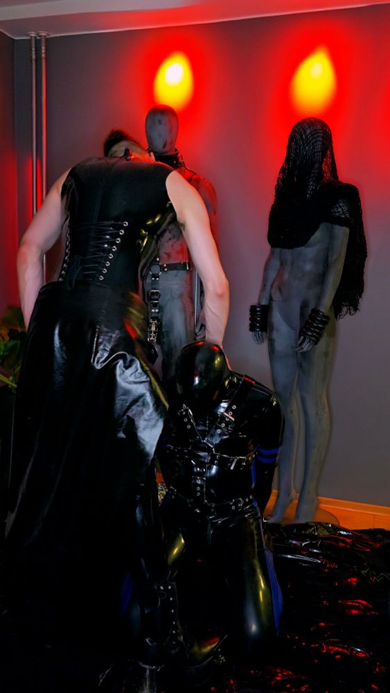 A Dominant in full latex is standing above a kneeling rubber gimp forcing its head downwards.
The background is dominated by two large mannequins in bondage gear.