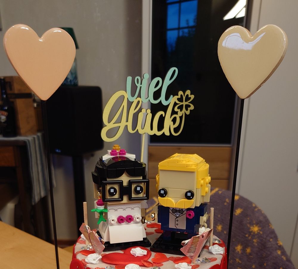 Ein Hochzeitspaar aus Lego steht auf der obersten Ebene einer Torte. Erkennbar auf der Torte sind Herzen aus Geldscheinen, aus Stoff und aus Keramik. Im Hintergrund steht viel Glück. Die Torte war das Hochzeitsgeschenk...