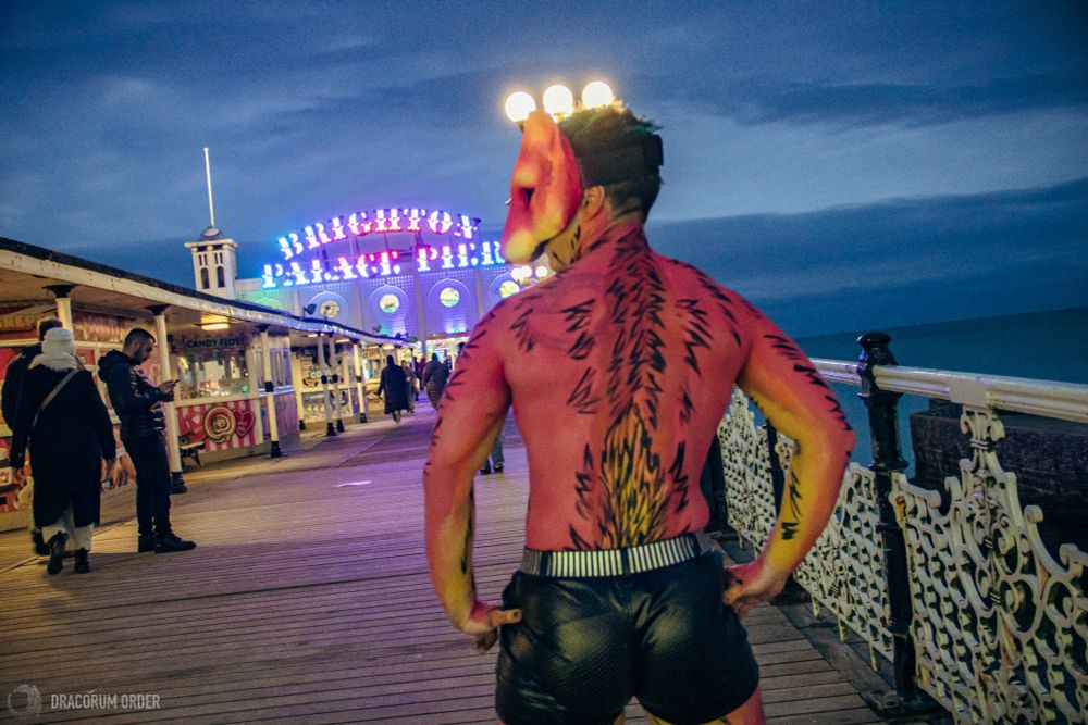 A photo of Alix wearing black shorts and their UV-reactive red and yellow weasel mask with body paint to match it. They are photographed from behind standing on the boardwalk at Brighton Pier in the dusk, with the Brighton Palace Pier sign illuminated ahead of them. Their hands are on their hips as they glance back over their shoulder.