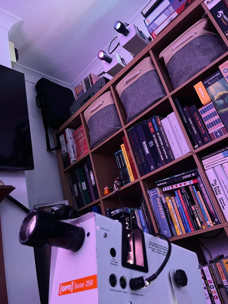 A Solar 250 projector in the foreground, with a large bookshelf in the background on which are sat two more. 