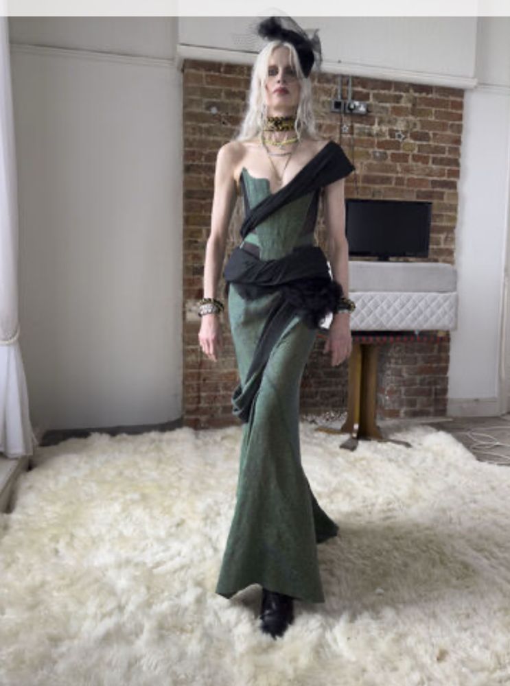 A blonde model in a white room with a brick fireplace and a fluffy white fur rug. She is wearing a dark green silk mermaid gown, with black accents and a black sash wrapping around one leg, around her waist, leading up to a strap across the opposite shoulder. She is also wearing black jewelry and a black tulle hat.