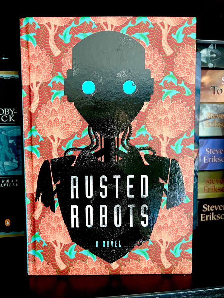 Underneath the dust jacket of the special edition hardcover of Death Of The Author by Nnedi Okorafor: a robot with circular blue eyes and wires coming out of its neck from Rusted Robots- the fictional book within a book from the story. 