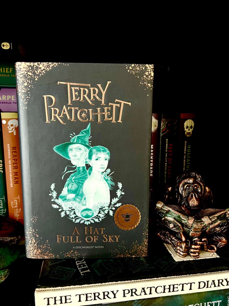 Tiffany Aching, witch of The Chalk, and Esmeralda Weatherwax, witch of Lancre, side by side on the book cover for the gift edition hardcover of A Hat Full Of Sky by Sir Terry Pratchett. 