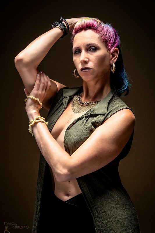 A Caucasian woman in a green vest and black pants. She has pink and blue hair, pulled back way from her face. She has slender, muscular arms, both with bracelets on them. Her arms are posed across her torso, one hand rests on her head. The vest is open, showing the line of her body from her neck to her midsection. Leigh Targaryen in the studio with FightGuy Photography