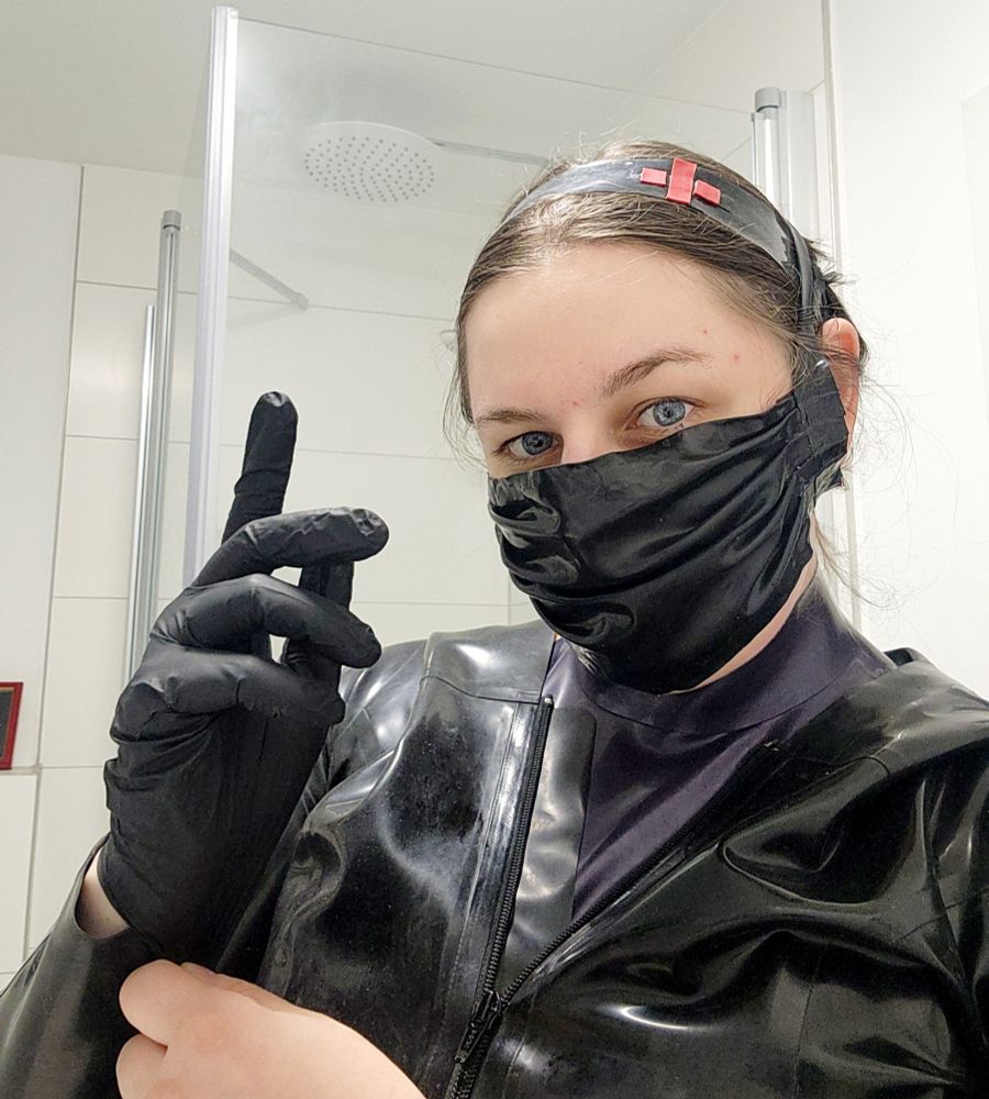 Black Latex nurse look with an open jacket, surgeon style mask, headband with a red cross. Putting on a latex glove and looking at you
