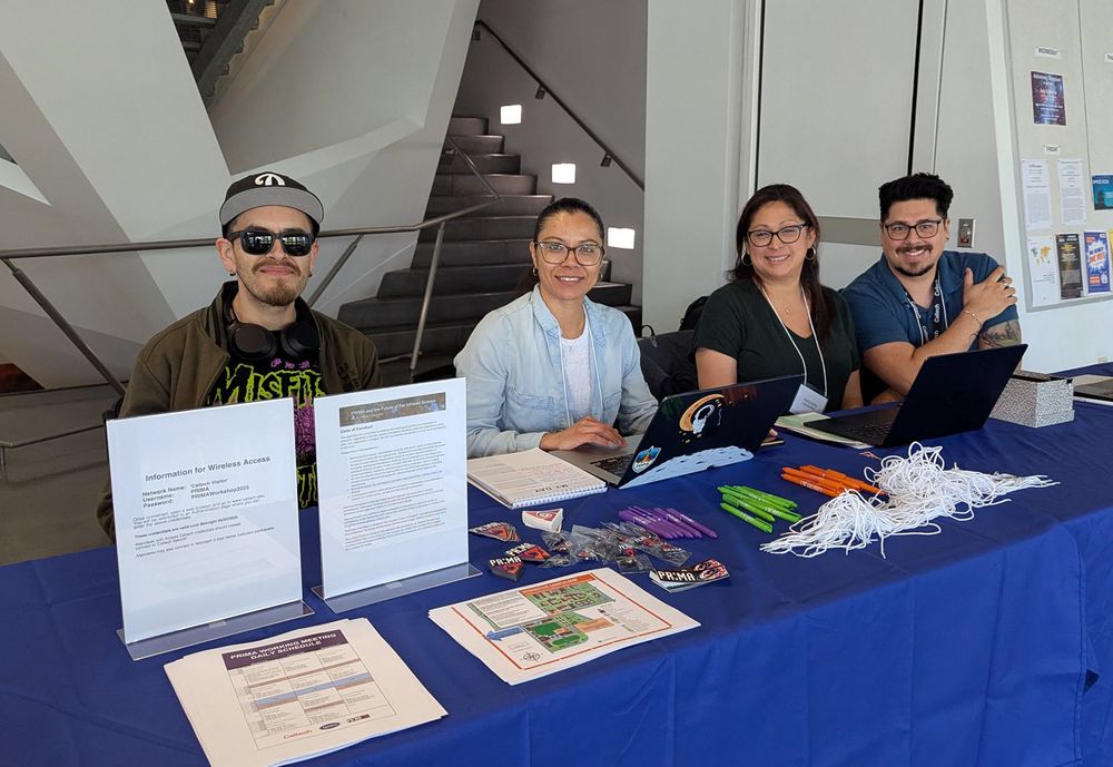 This is a group photo of four people sitting behind the registration table at the PRIMA working meeting, held in the Cahill Center for Astronomy and Astrophysics on the Caltech campus. The table holds brochures, lanyards, and swag (pens, stickers, magnets, etc.) for event attendees. From left to right: Daniel "Pina" Muro, Teresa Molano, Nancy Solis, and Sean Kindt. Image credit: Marcy Harbut, Caltech/IPAC