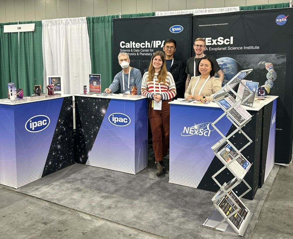 This is an image of the combined Caltech/IPAC and NExScI booth at the 246th meeting of the American Astronomical Society (AAS) held in Anchorage, Alaska from June 8 to 12, 2025. 

Visual description: Five people -- three adult men and two adult women -- stand behind two waist-level tables branded with the IPAC and NExScI logos. Behind them are banners that say Caltech/IPAC and NExScI. The image was taken in the Exhibit Hall area of the convention center in Anchorage, Alaska where the science meeting was held from June 8 through June 12, 2025. 