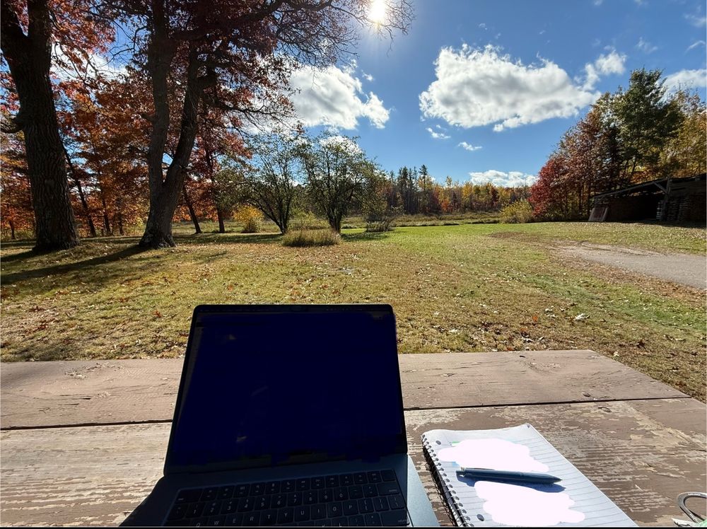 Picture taken in the middle of the day, outside, looking at a laptop and a notebook on a worn brown picnic table top. Just past that, a fading green yard of grass and brown fallen leaves, with some random trees in the middle and background starting to turn fall colors. The sky is blue with bright fluffy white clouds.