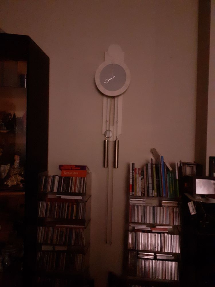 Visible between two half-tall and heavily laden shelving units is a wall-mounted pendulum clock, with the weights half-way down the wall.