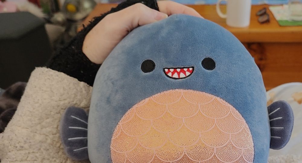 White hand in black fingerless glove resting on top of Shark plushie - a grey squishmallow with big black eyes, small red mouth, white teeth, pink reflective belly scales, small fins on either side.