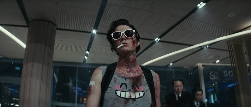 Photo from the movie Kate where she is in an office with a blood-stained smiley face singlet, cigarette dangling from her mouth and dark sunglasses over a bloodied face.