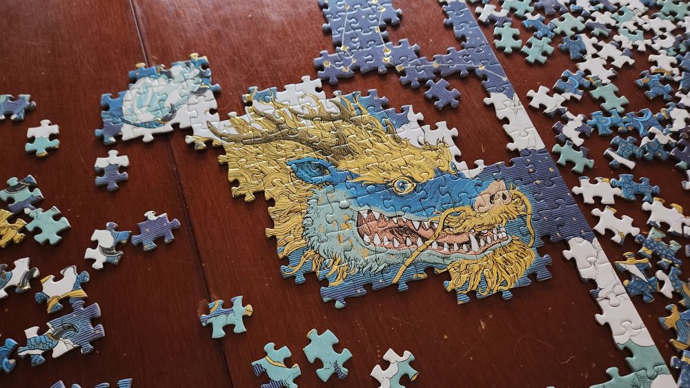 Close-up of dragon head in a partly completed blue and yellow dragon jigsaw with gold highlights, on a red-brown wooden table.