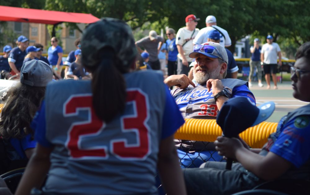 Dan Ferreira, coach of the Chicago Junior Wheelchair Cubs and adaptive sports and event coordinator with the Chicago Park District, talks to his team, who are all sitting in sport chairs and wearing grey jerseys with blue sleeves and red lettering.