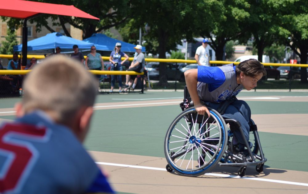 Lex McGuire, a pitcher and mentor on the Chicago Junior Wheelchair Cubs wearing a grey jersey with blue sleeves and red lettering,  rushes to first base during a skill challenge as teammate Cooper Roberts watches during the 2025 Wheelchair Softball Junior World Series at California Park's paved baseball diamond on Saturday.