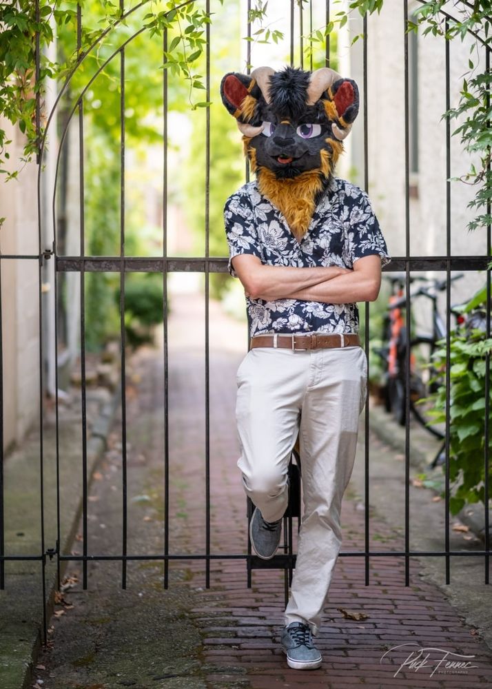 A man wearing a ram fursuit head, leaning against a gate