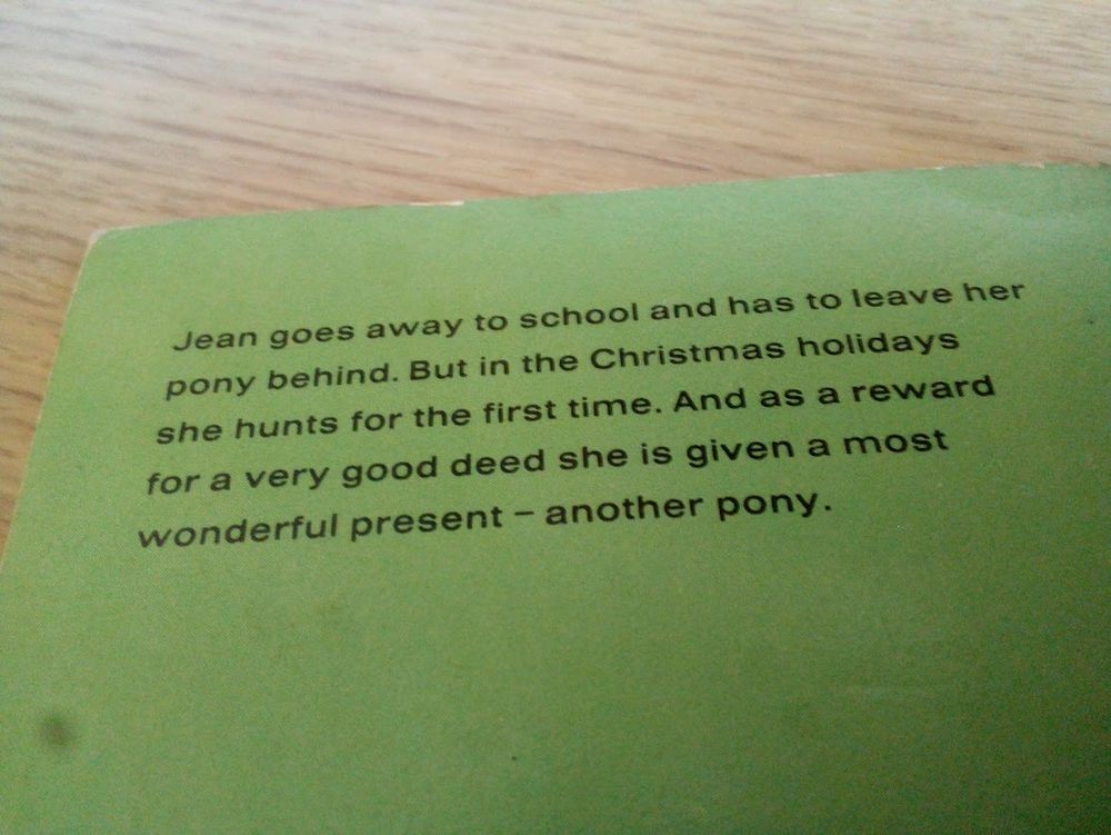 A piece of blurb on the back of a book. The book is green and sits on a brown desk. The blurb reads: "Jean goes away to school and has to leave her pony behind. But in the Christmas holidays she hunts for the first time. And as a reward for a very good deed she is given a most wonderful present - another pony."