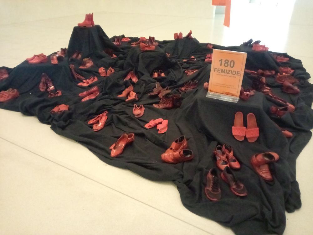 Ein schwarzes Tuch auf dem Boden darauf rot eingefärbte Schuhe und ein Schild 180 Femizide im Jahr 2024