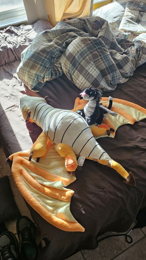 The yellow plush lies upside down on my bed, exposing its belly while the little black plush gives it belly rubs